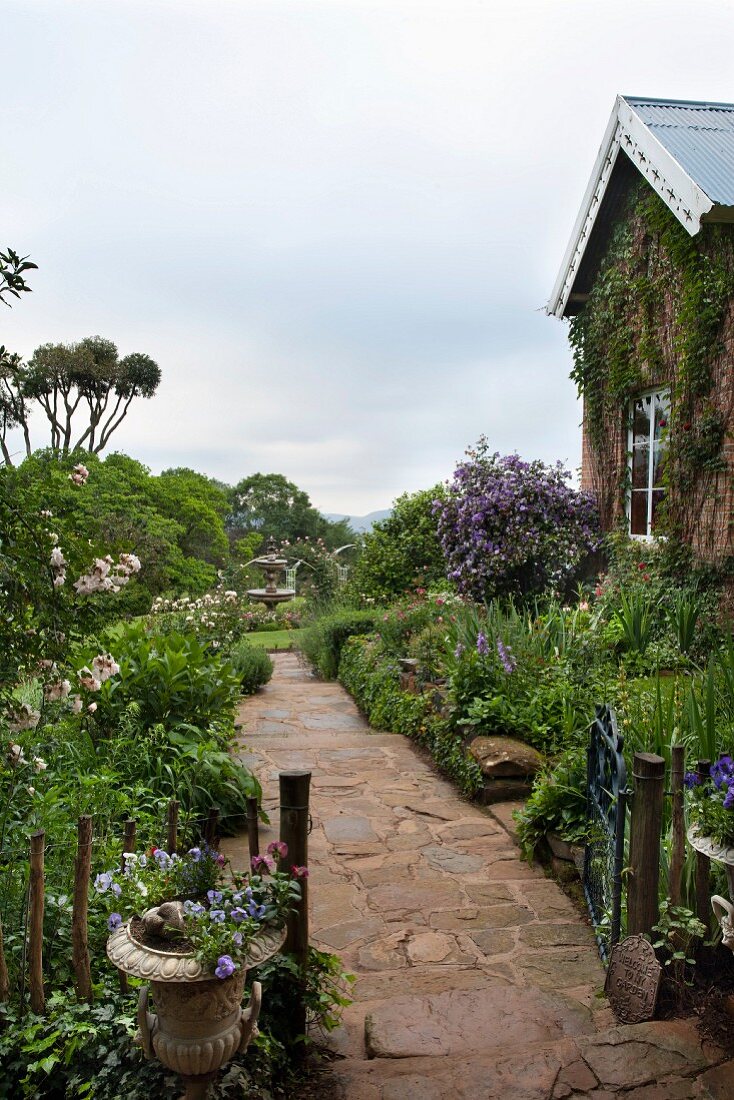 Schmaler Weg mit Natursteinbelag in blühendem Garten, seitlich teilweise sichtbares Wohnhaus