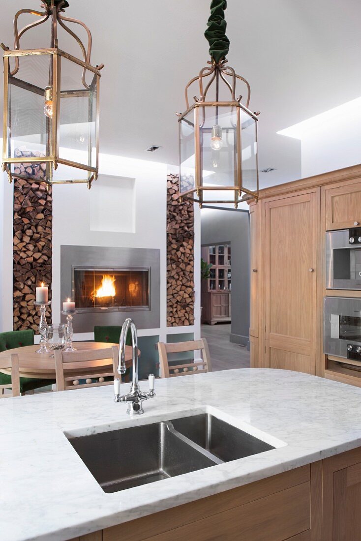 Kitchen island with pale marble top and integrated sink below lantern-style pendant lamps; dining area with fire in fireplace in background