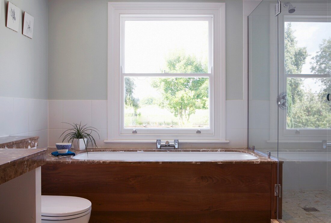 Eingebaute Badewanne mit Holzfront vor Fenster, seitlich Glaswand vor Dusche
