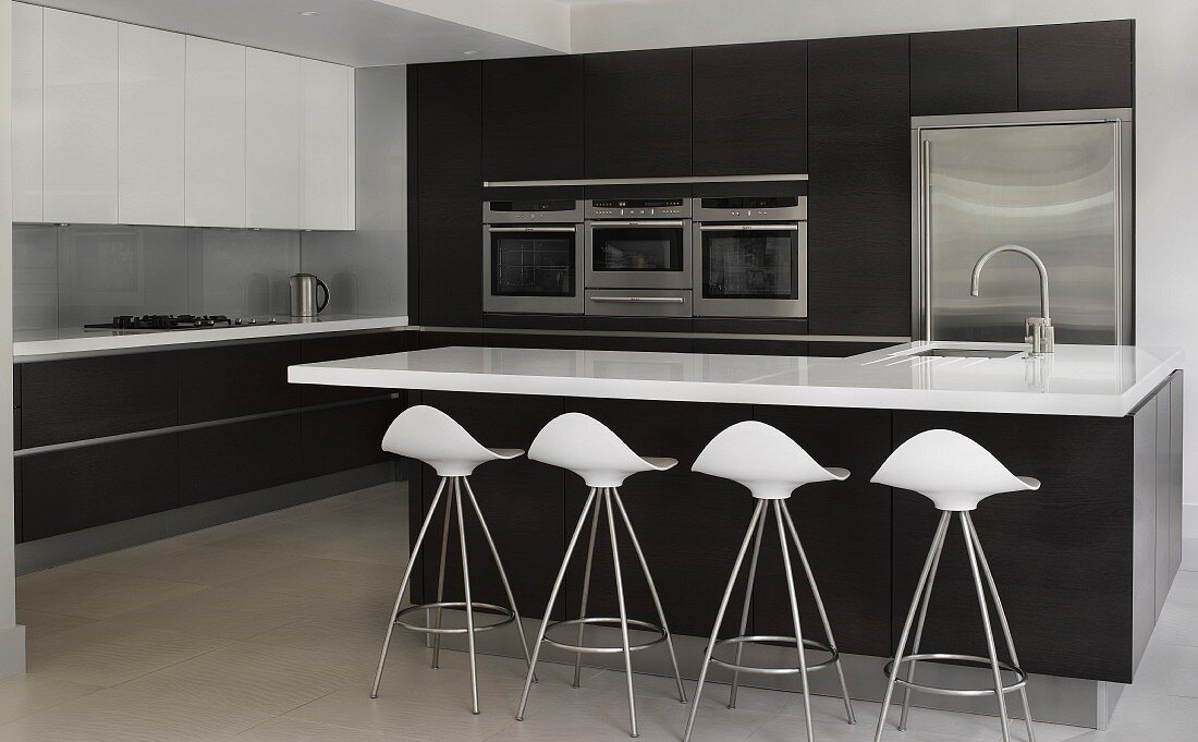 Designerbarhocker mit weissen Sitzschalen vor Kücheninsel mit schwarzem Unterbau, im Hintergrund Einbauschrank mit Küchengeräten