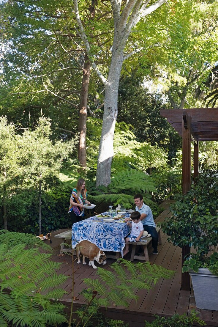 Familie mit Kind und Hund auf Holzterrasse, gedecktem Tisch, umgeben von tropischen Pflanzen