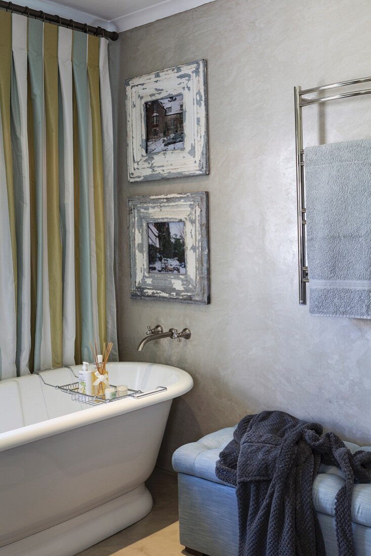 Pictures in vintage frames above retro bathtub next to upholstered bench and heated towel rack on marbled wall