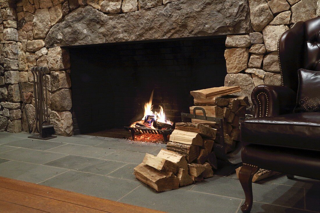 Open fire in rustic interior with stone wall, stack of firewood and restored leather armchair to one side