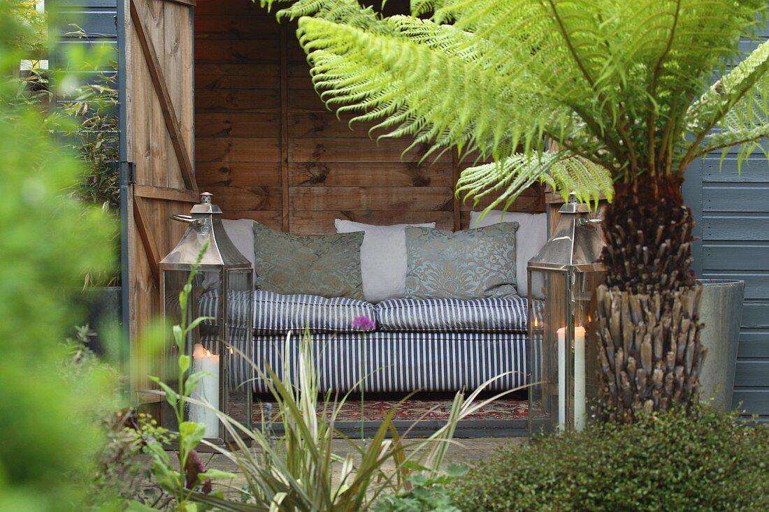 Sofa in wooden shed, two lanterns and tree fern in garden
