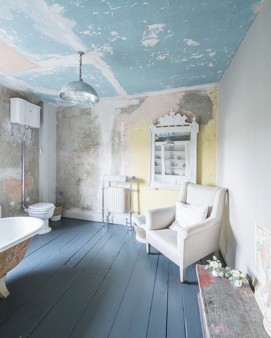 Armchair next to antique shelving, toilet and free-standing bathtub in large bathroom with patinated walls and ceiling