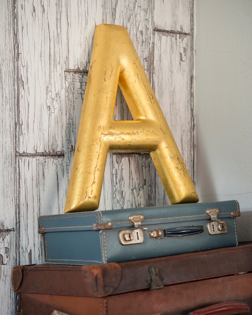 Gilt decorative letter A on stack of vintage suitcases leaning against peeling board wall