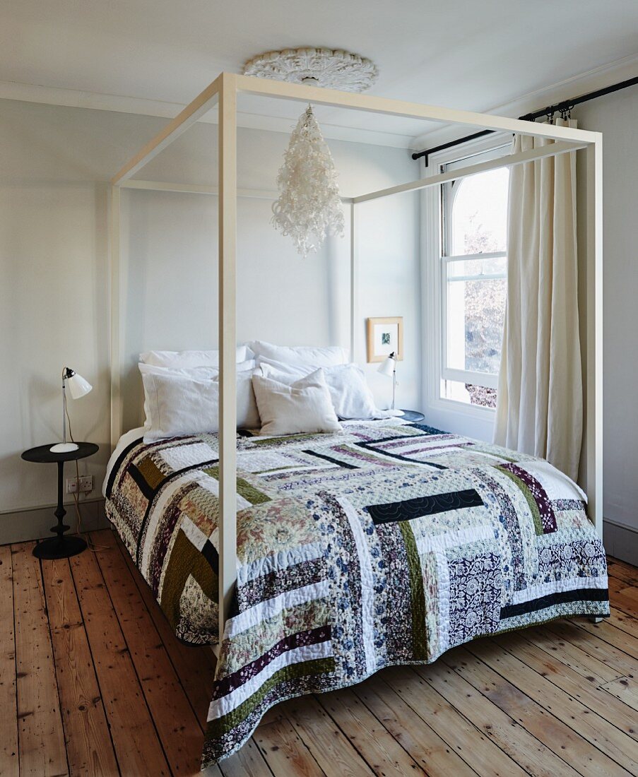 Patchwork cover on double bed with white-painted canopy frame in simple bedroom with wooden floor