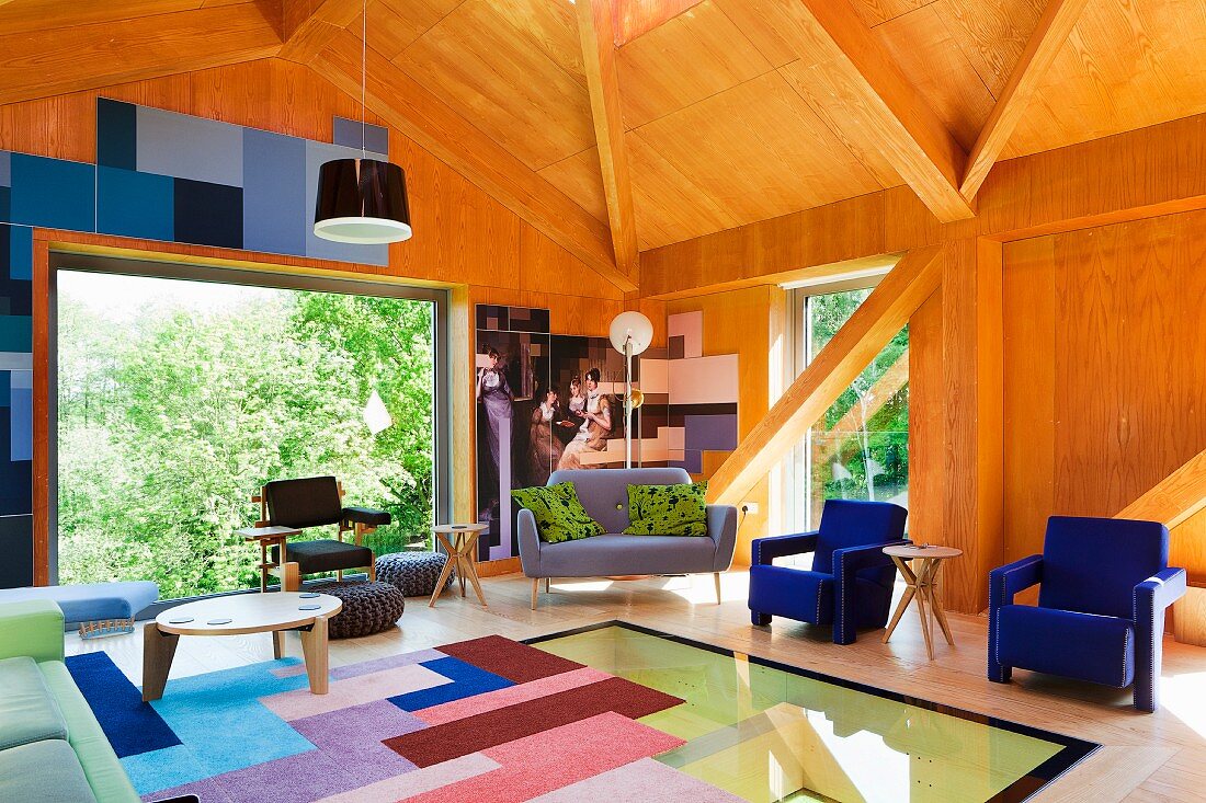 Patchwork rug on glass floor and blue armchairs in wood-panelled interior of contemporary building