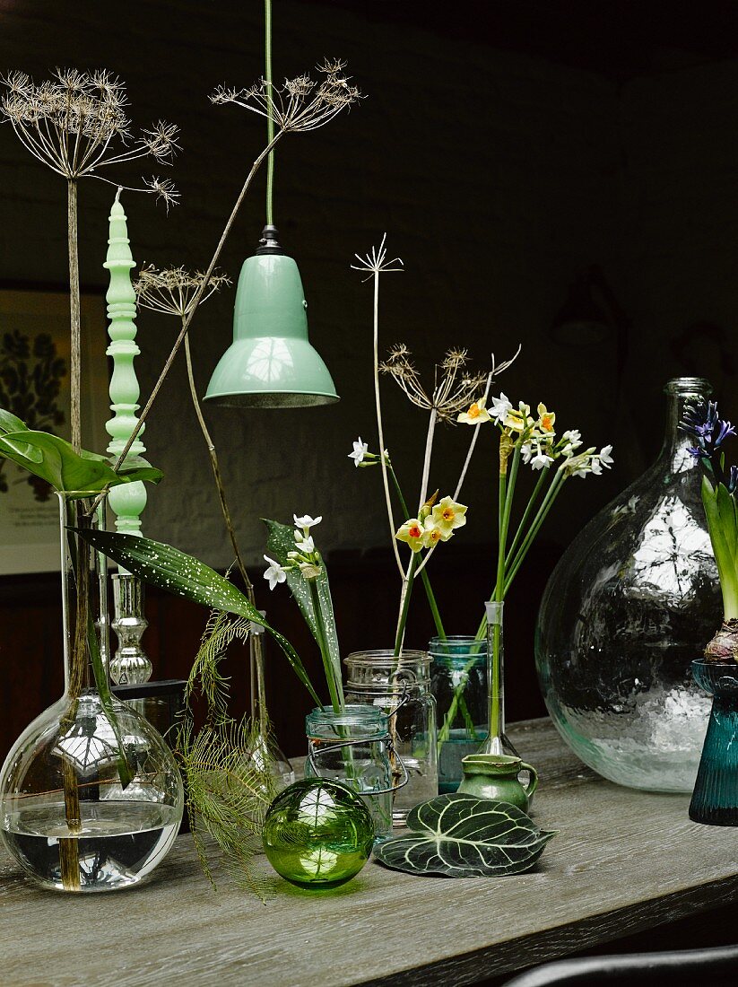 Various flowers in vases and turned candle in candlestick next to vintage, green pendant lamp