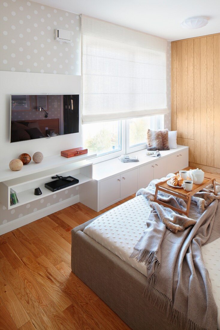 Double bed opposite fitted cabinets below window, wall mounted shelves and flatscreen TV in modern bedroom