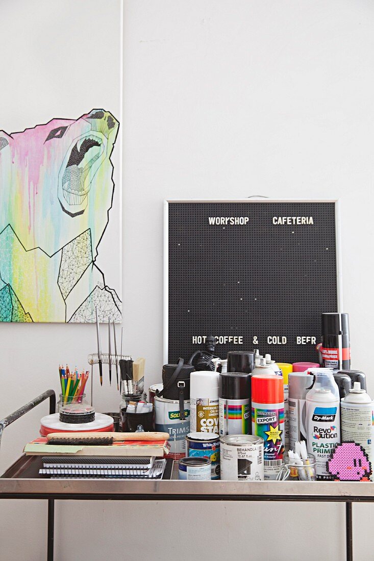 Paint pots, spray cans and drawing utensils on metal table in front of picture of bear and pegboard