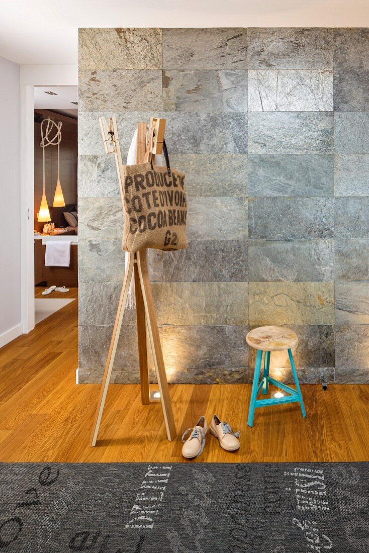 Bag hanging from wooden coat stand, stool and rug with pattern of lettering in front of wall covered in stone tiles
