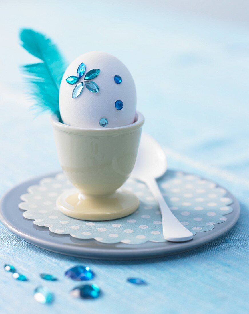 Weisses Hühnerei verziert mit blauen Glitzersteinen & Feder im Eierbecher