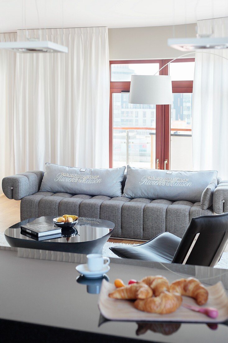 Designer lamps above grey couch in elegant living room; croissants on breakfast bar in foreground