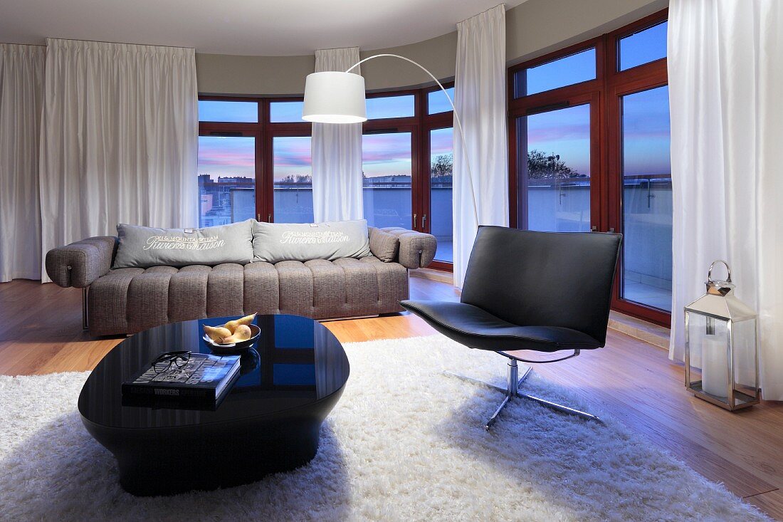 Eleganter Sitzbereich mit Ledersessel und Hochglanztisch vor geschwungener Fensterfront in der Abenddämmerung