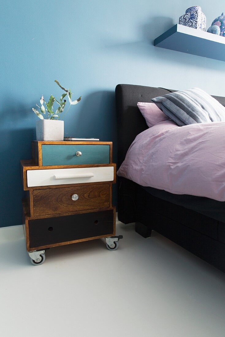 Bedside cabinet made from different drawers on castors next to bed against blue-painted wall