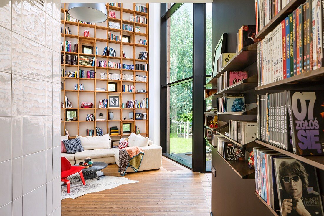 Seitlich Regal mit Büchern, im Hintergrund Loungebereich mit hellem Polstersofa vor raumhohem Bücherregal aus Holz in zeitgenössischem Wohnhaus