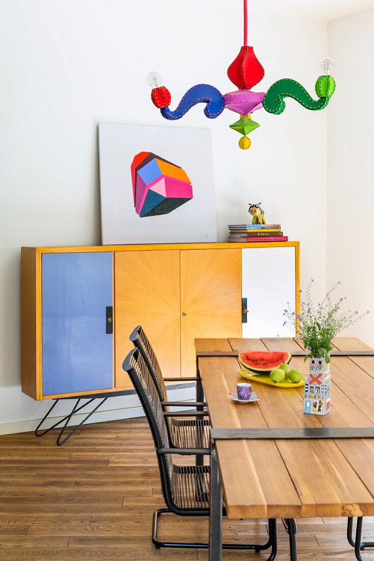 Postmodern pendant lamp with colourful arms above dining table with wooden top mounted on metal frame, metal armchairs and sideboard with colourful doors in background