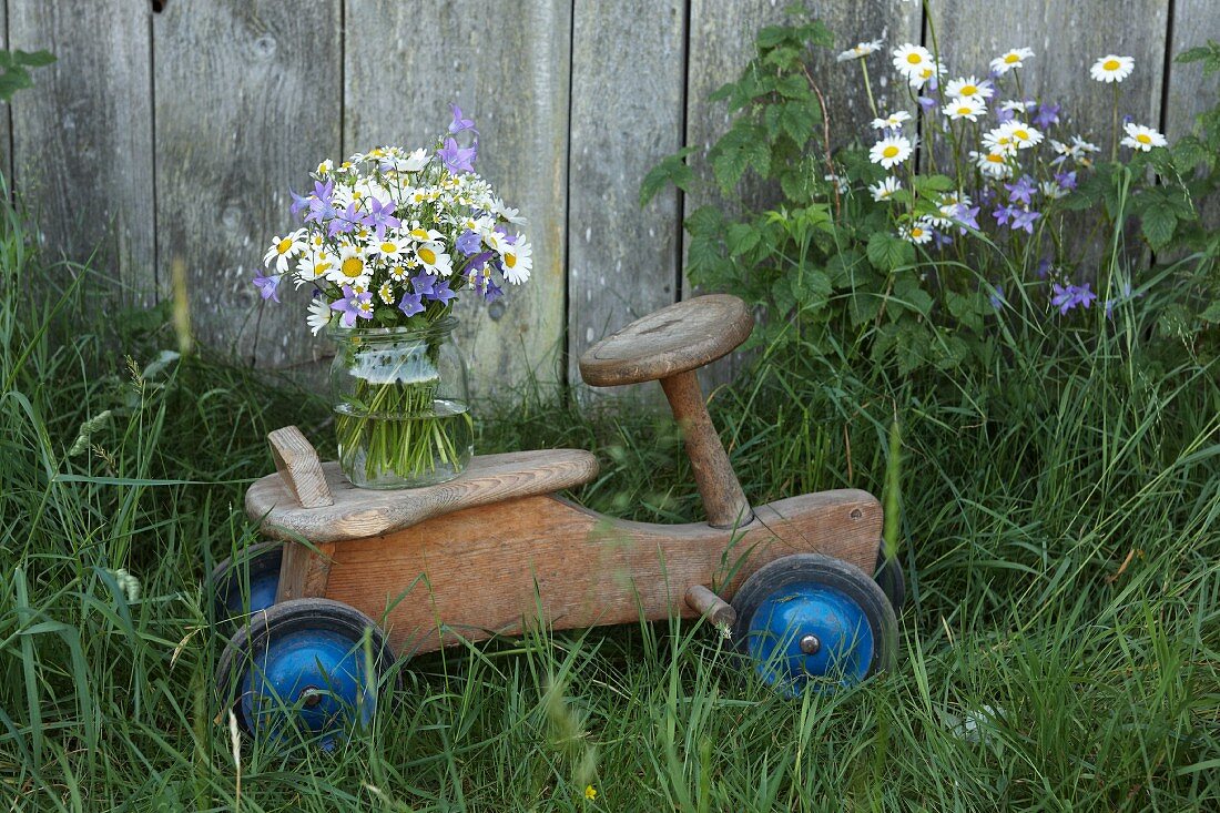 Glockenblumen, Margeriten und Kamillenblüten im glas auf rustikalem Kinderfahrzeug aus Holz