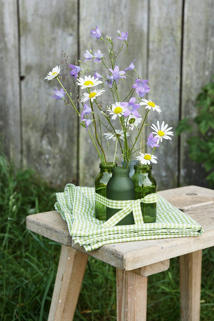 Zarte Glockenblumen und Margeritenblüten in grünen Flaschenvasen und grün-weiss karierte Tischdecke auf rustikalem Holzschemel