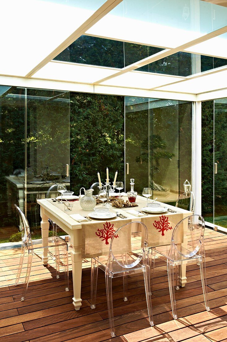 Ghost Stühle um gedeckten Tisch auf Holzterrasse eines verglasten Wintergartens, oberhalb abgehängte Decke mit opakem Glas