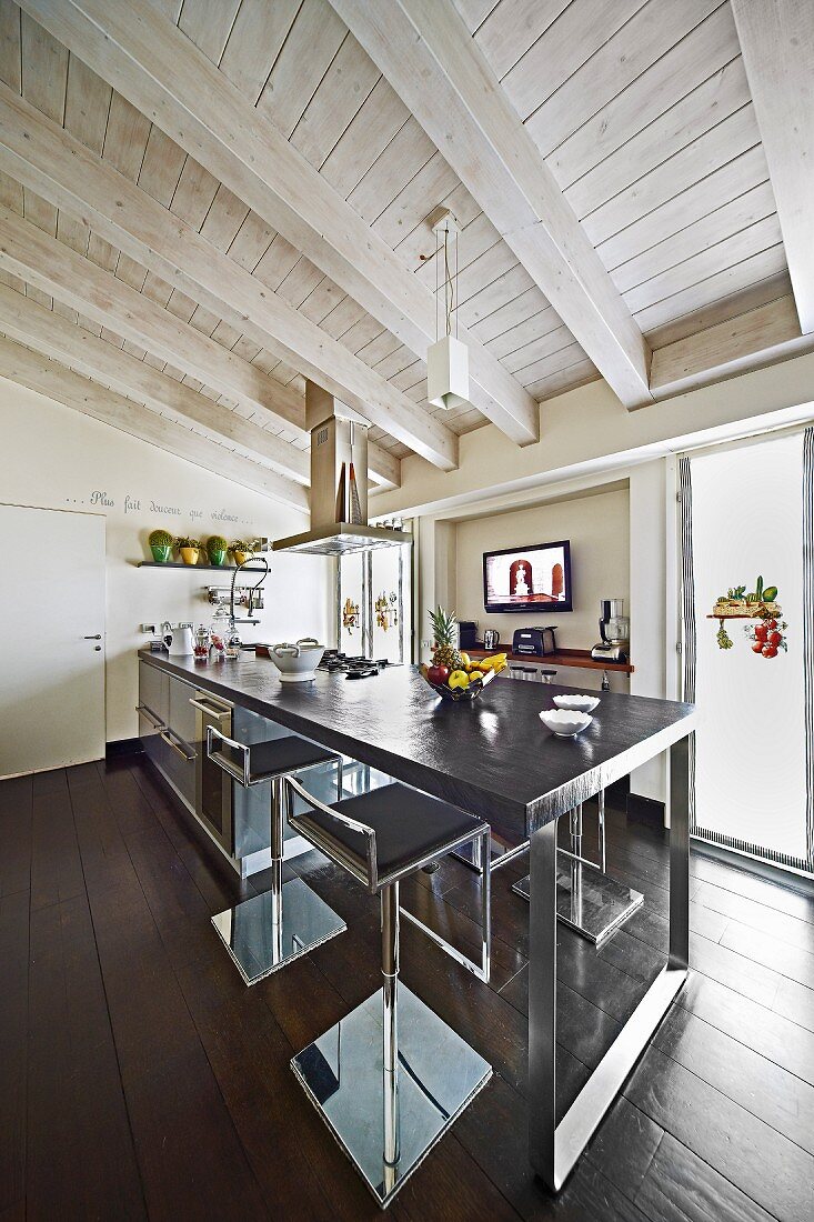 Moderne Küchentheke mit dunkler Arbeitsplatte auf Metallgestell und Barhocker in ländlichem Wohnraum mit weiss lasierter Holzdecke