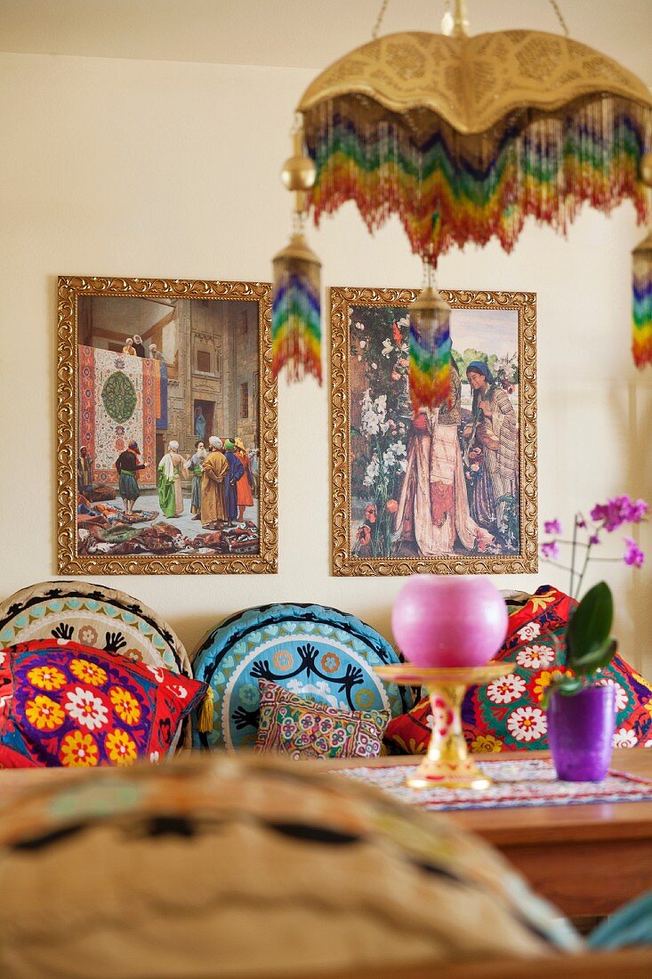 Verschiedene bunt gemusterte Kissen auf einer Bank am Tisch, darüber zwei orientalische Bilder