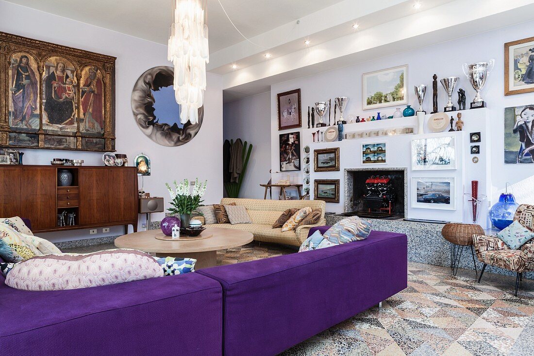 Violettes Sofa um runden Couchtisch in modernem Wohnraum, an Wand gerahmte Bilder und Pokalsammlung