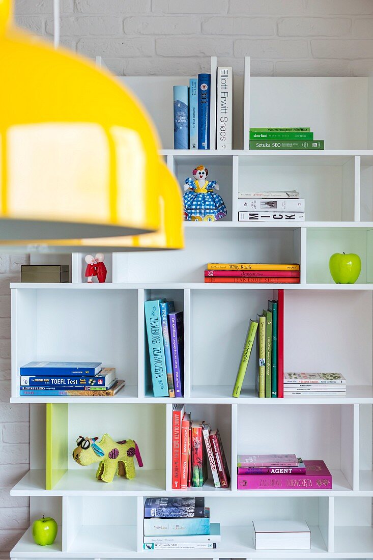 Weisses Regal mit Büchern und bunten Figürchen; im Vordergrund gelbe Pendelleuchten