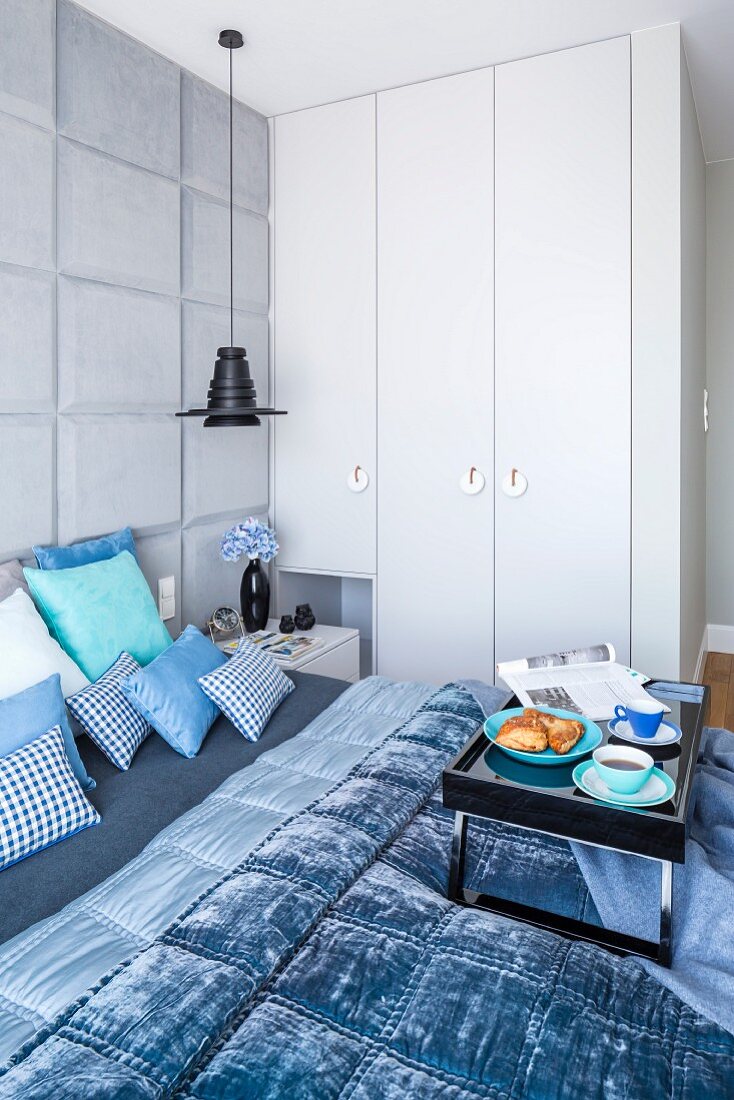 Frühstückstablett auf Doppelbett mit Kissen und Decken in Blautönen in Schlafzimmer mit Einbauschrank
