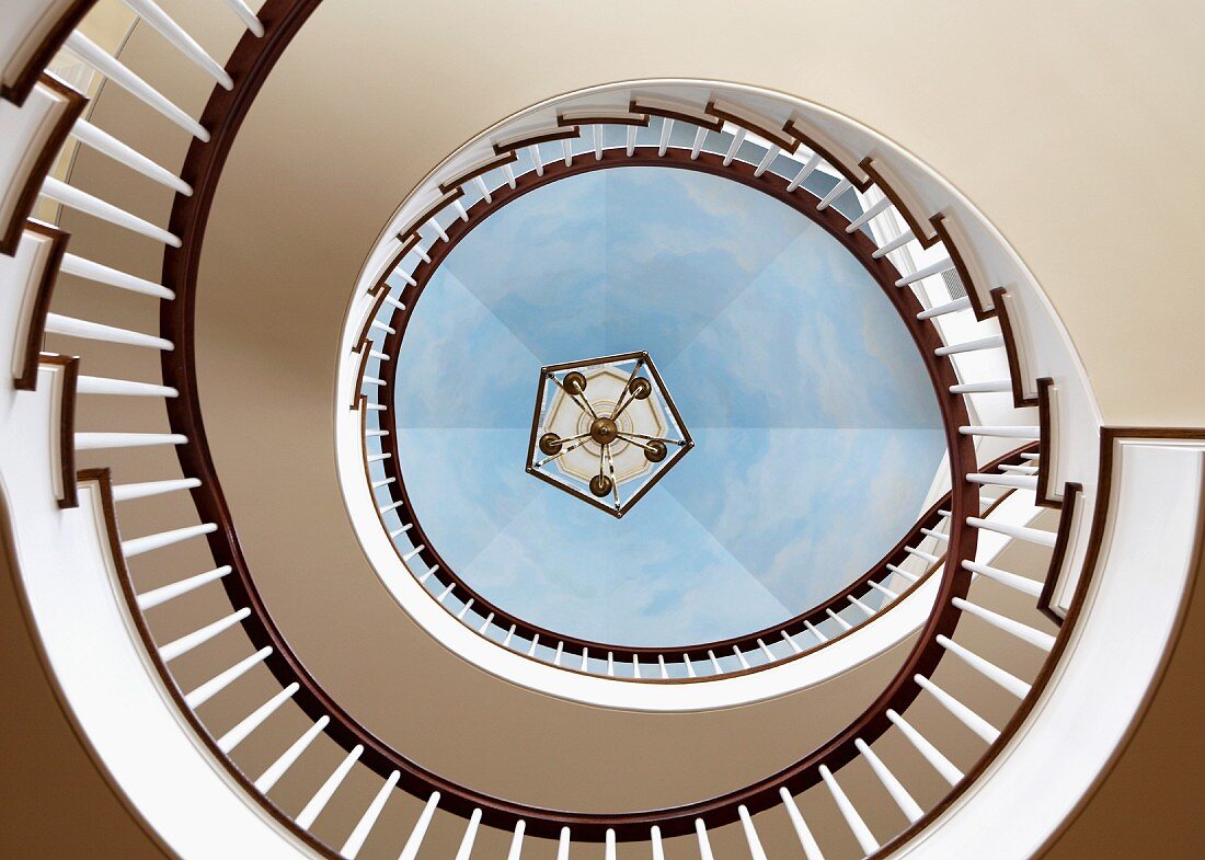 View up through stairwell of elegant, winding staircase with trompe l'oeil ceiling painted to resemble sky
