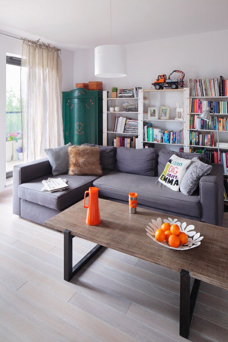 Coffeetable mit orangefarbenen Accessoires, grauviolette Eckcouch und einfaches Bücherregal im Hintergrund