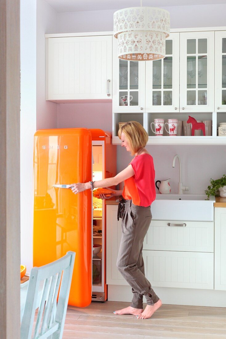 Frau in weisser Landhausküche an orangefarbenem Retro-Kühlschrank