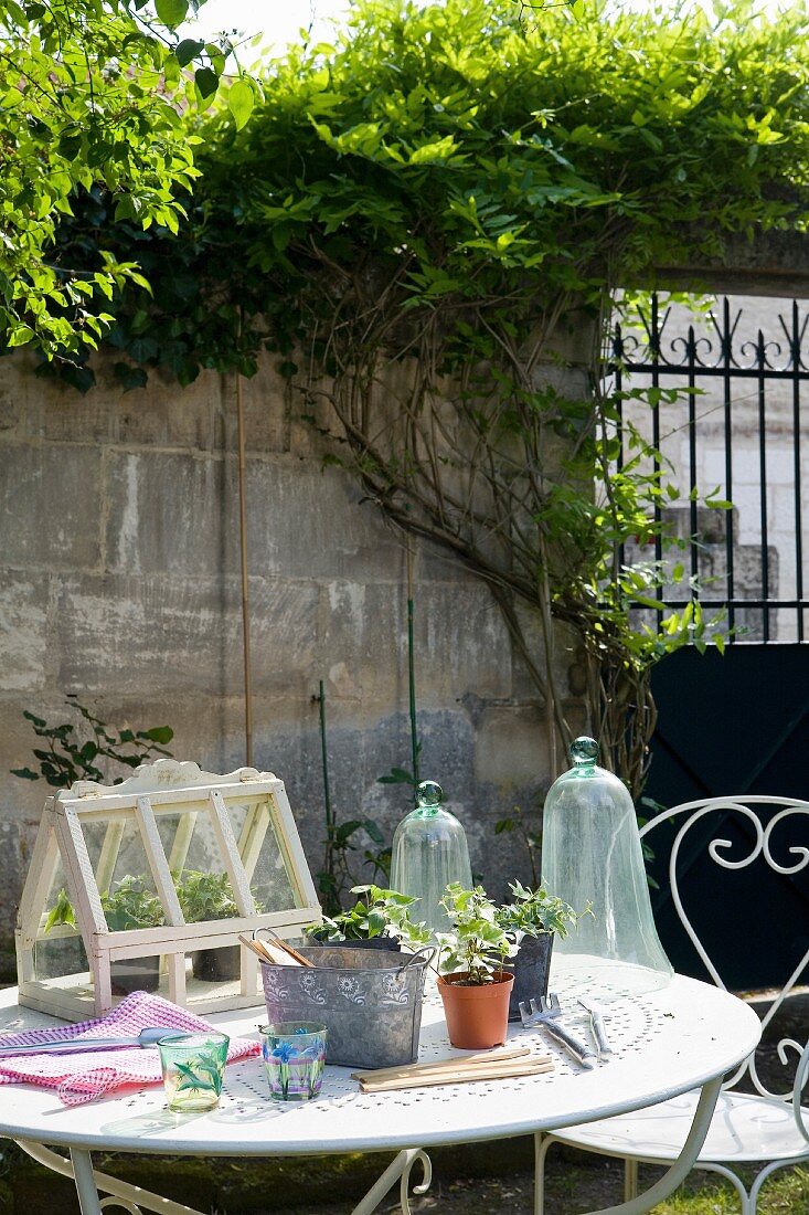Ummauerter Garten mit verschnörkelten Gartenmöbeln aus Metall, auf dem Tisch ist ein Minigewächshaus und Werkzeug