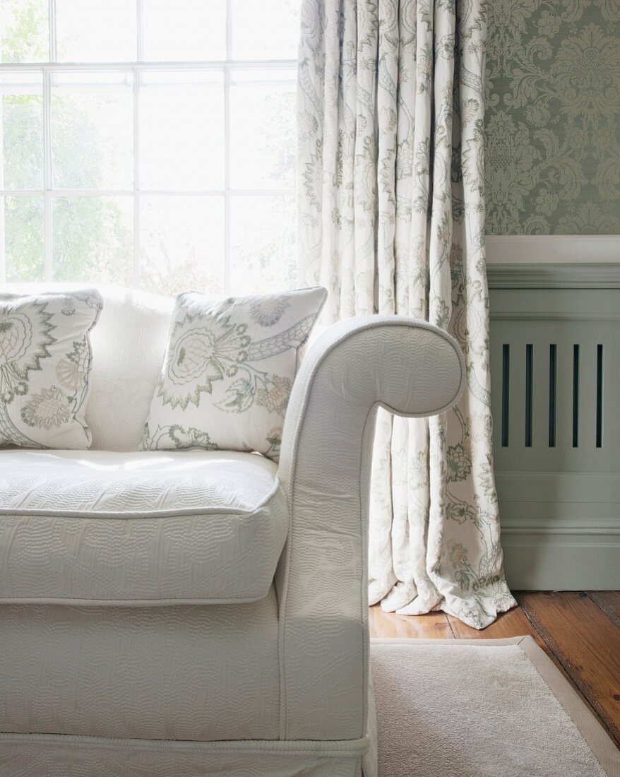 Ecke eines klassischen Sofas vor Sprossenfenster mit Vorhang und Ornament-Tapete