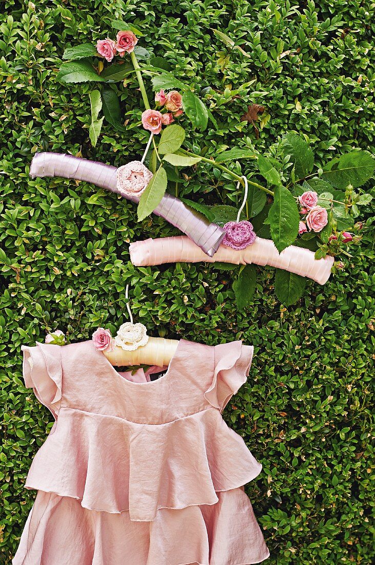 Pink, ruffled dress and romantic coat hangers with crocheted flowers hanging from rose branch in box hedge