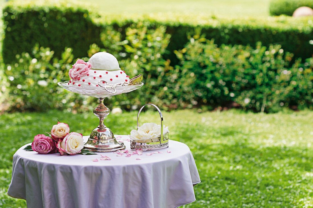 Romantic, festive cake on elegant cake stand and silver basket decorated with roses in summer garden