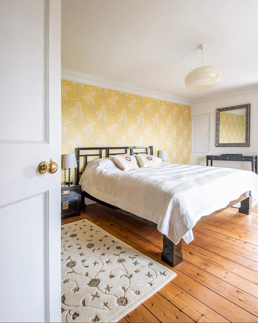 Blick durch offene Tür auf Doppelbett vor tapezierter Wand in schlichtem Elternschlafzimmer mit rustikalem Dielenboden