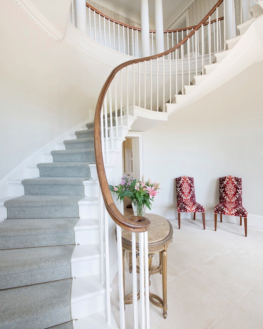 Curved staircase with pale grey runner and chairs with patterned upholstery in elegant foyer