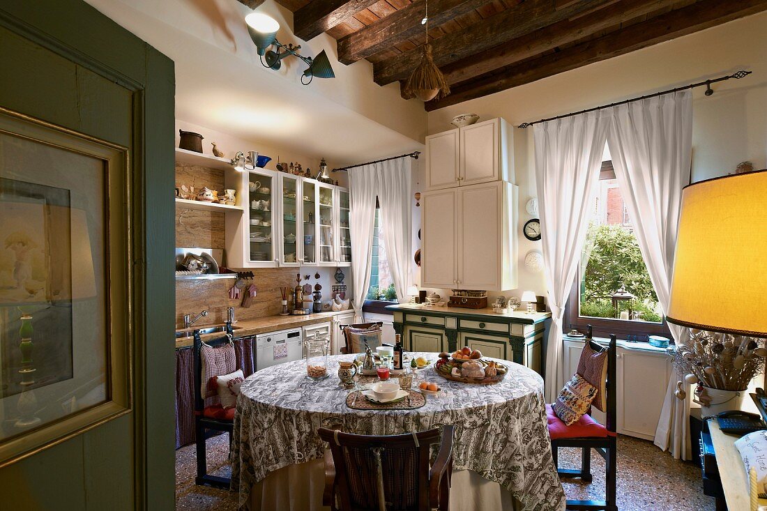Runder Tisch mit Tischdecke in mediterranerer Wohnküche, an Fenster geraffte, weiße Vorhänge
