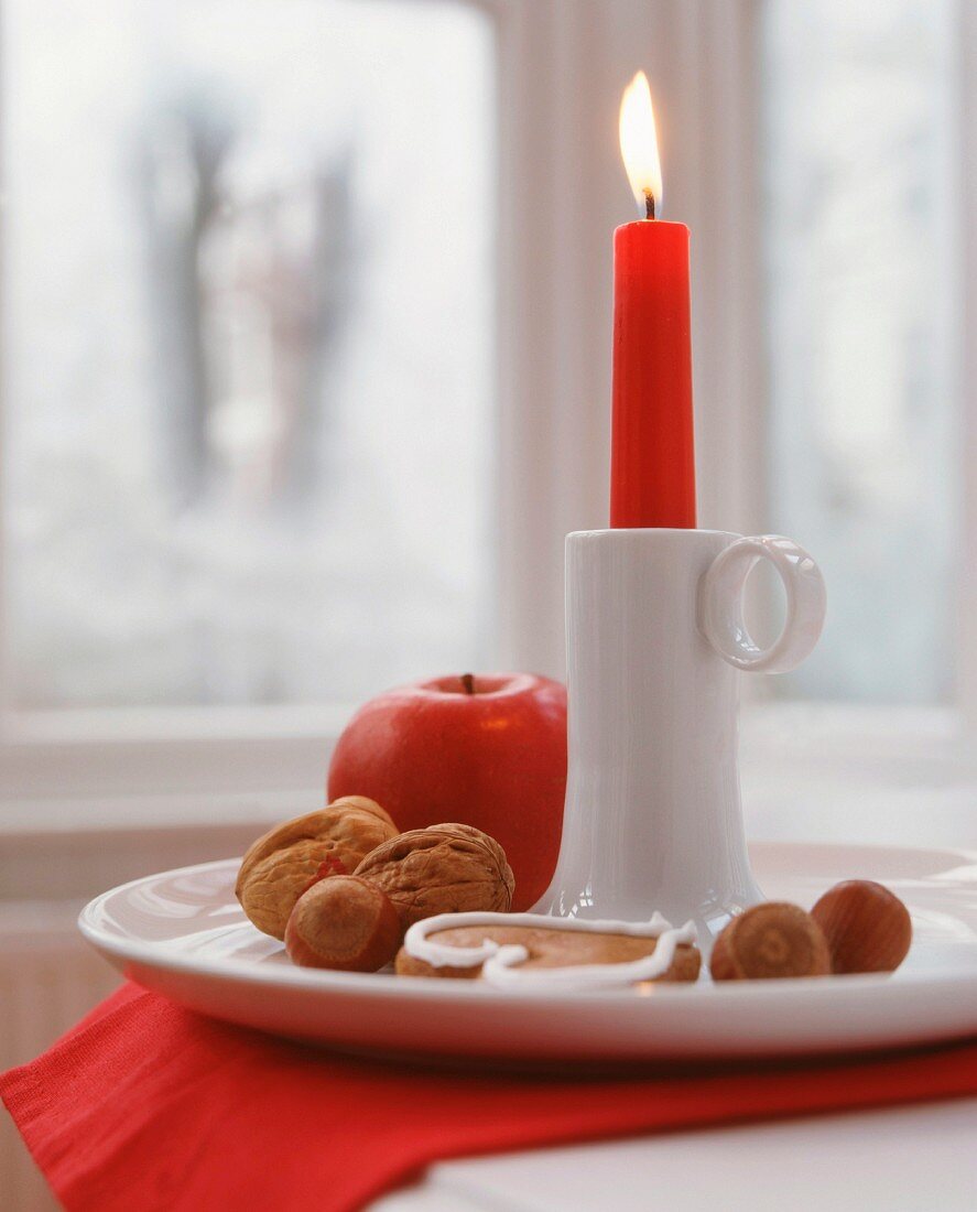 Lit red candle in white china candlestick with nuts, apples and gingerbread