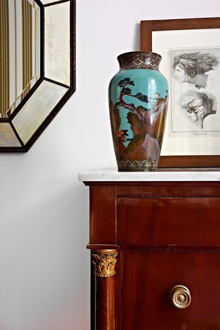 Bemalte, antike Vase auf Kommode, im Hintergrund gerahmte Zeichnung an Wand