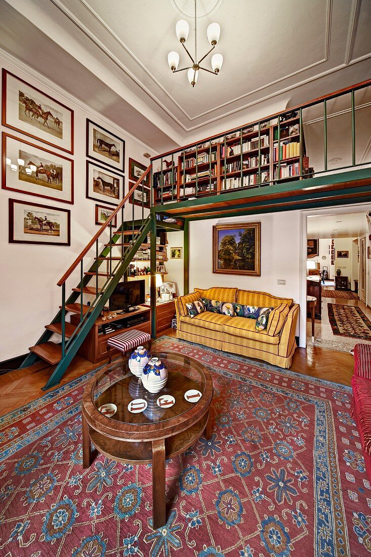 Antiker, runder Couchtisch auf gemustertem Teppich in offenem Wohnraum, im Hintergrund Treppe und Galerie in traditionellem Ambiente