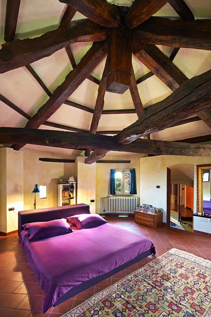 Double bed with purple bed linen in large bedroom below circular ceiling with old, massive wooden roof beams