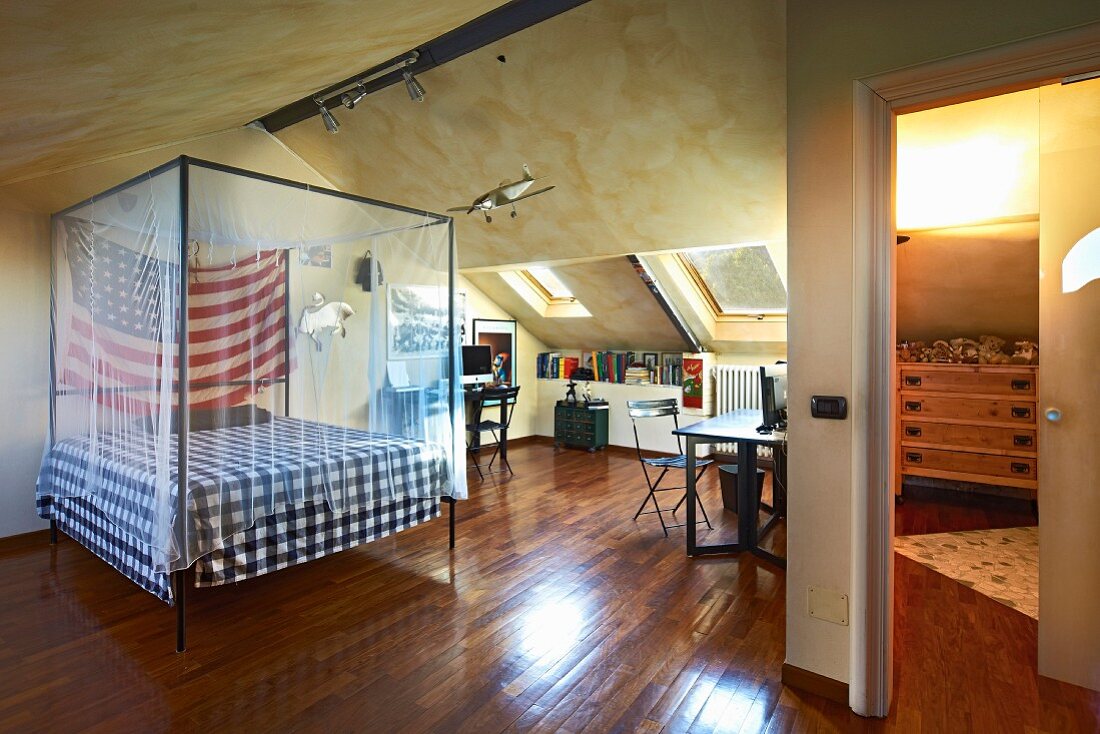 Bed with mosquito-net canopy and America flag in attic room with writing desk under sloping skylights