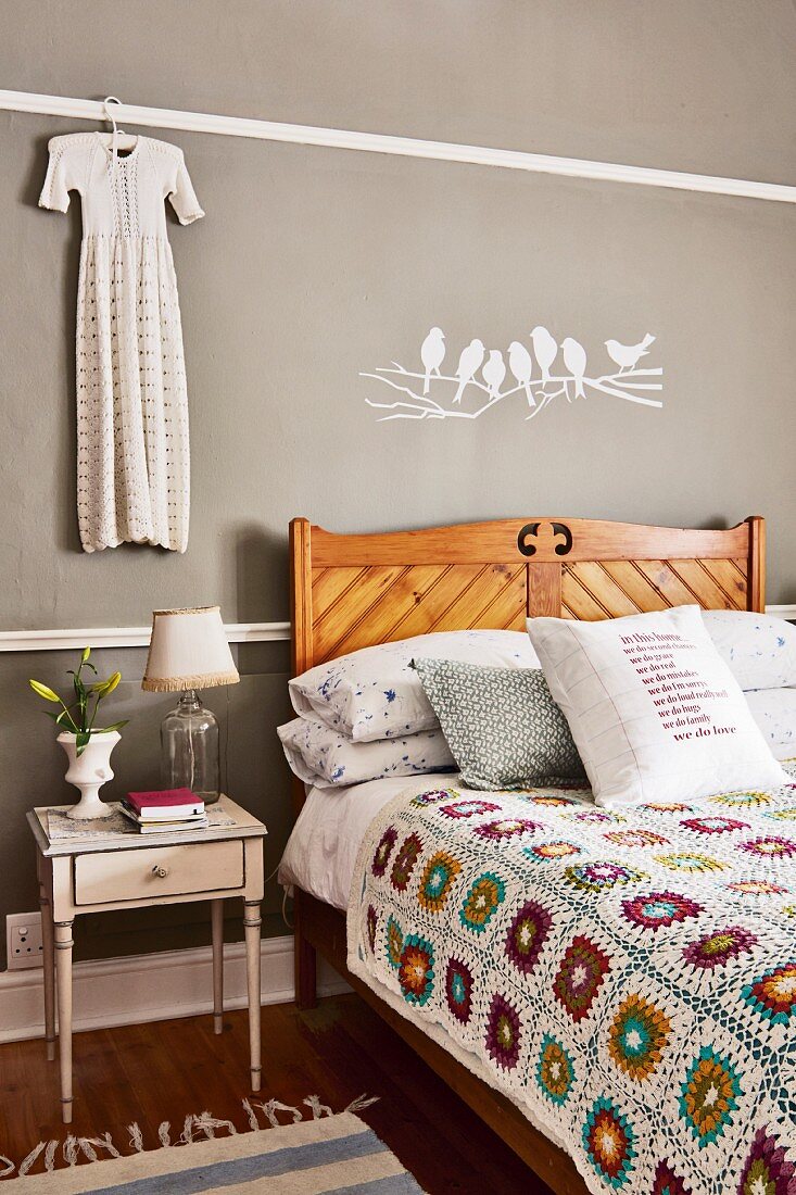 Crocheted patchwork blanket on wooden bed and antique bedside table below white bird motif on grey wall