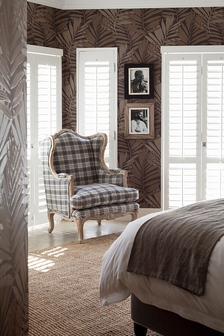 Wing chair in checked pattern in front of window with blinds and wall with leaf motif wallpaper