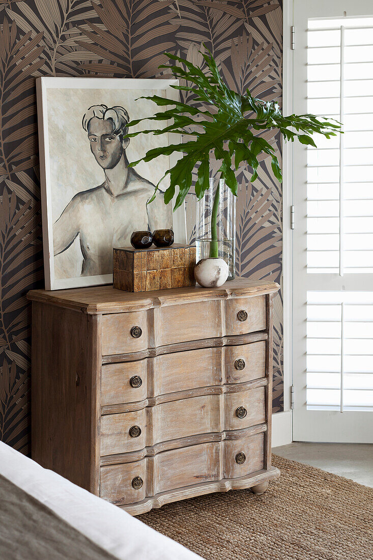 Wooden chest of drawers with plant and artwork in room with patterned wallpaper
