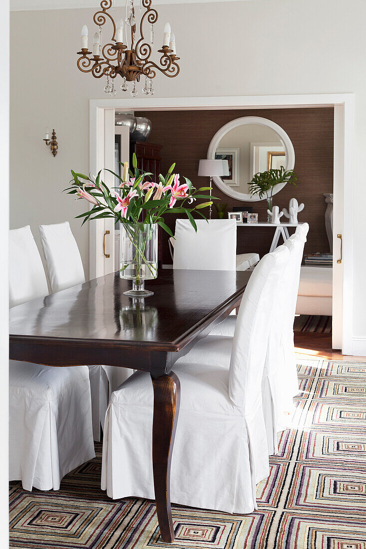 Classic dining room with wooden table, white chairs, chandelier and vase with fresh lilies