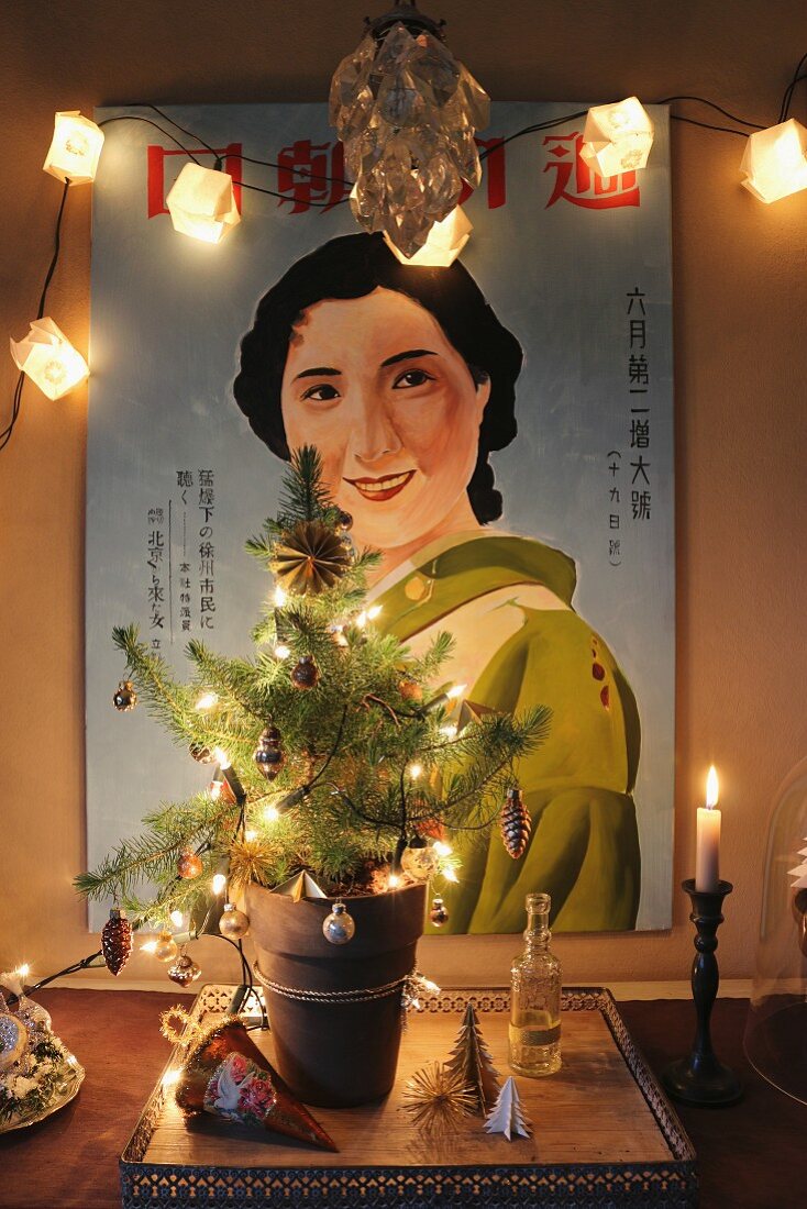 Geschmückter Weihnachtsbaum im Blumentopf mit Lichterkette vor beleuchtetem Bild mit japanischer Geisha